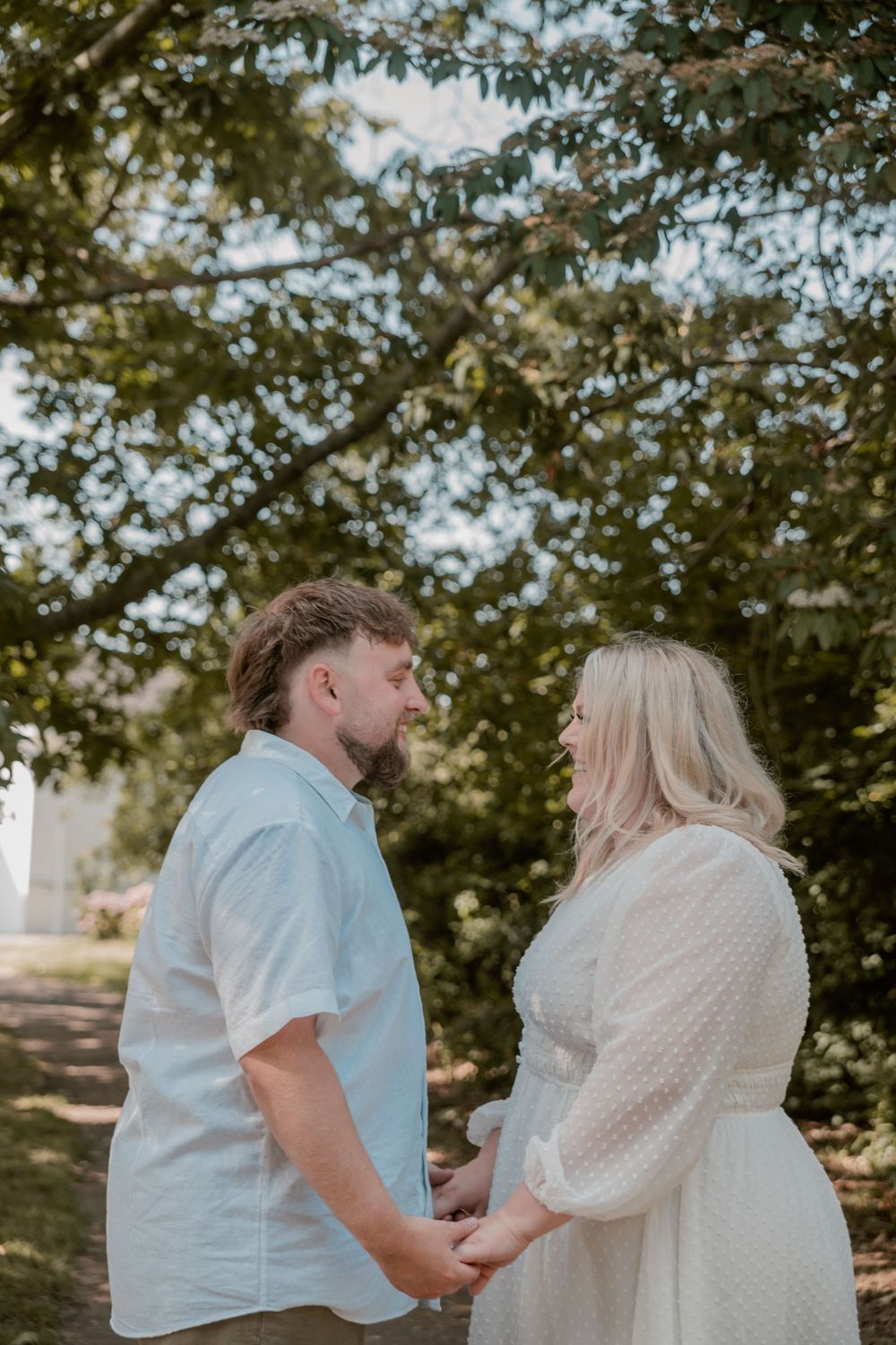 A couple holds hands facing each other under lush green trees, dressed in white and light blue outfits.