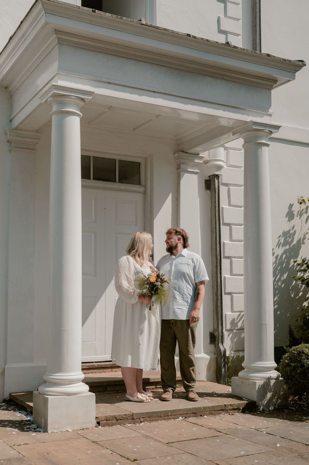 Bride and groom stand before white columned building entrance, she holds wildflower bouquet in white dress, he wears casual shirt.