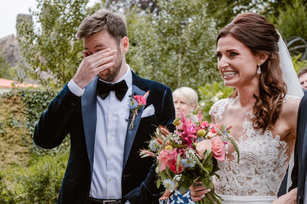 Emotional groom wiping tears beside smiling bride holding colorful bouquet at outdoor wedding ceremony.