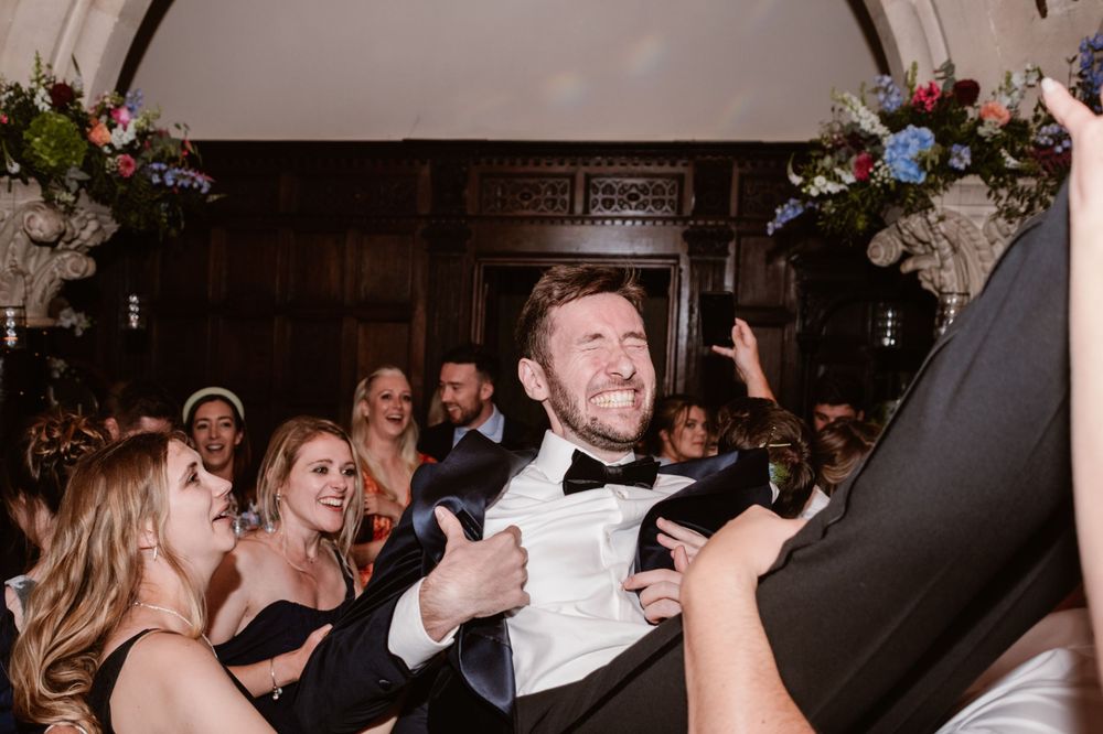 Groom in tuxedo laughing joyfully while being lifted by wedding guests during lively reception celebration.
