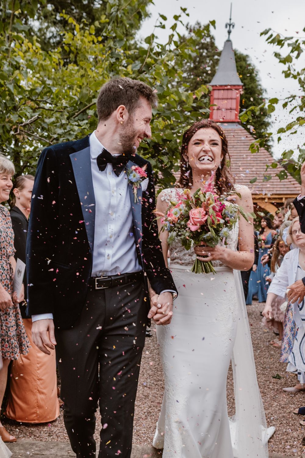 Bride and groom walk through colorful confetti shower, smiling joyfully, as guests celebrate their outdoor wedding ceremony.
