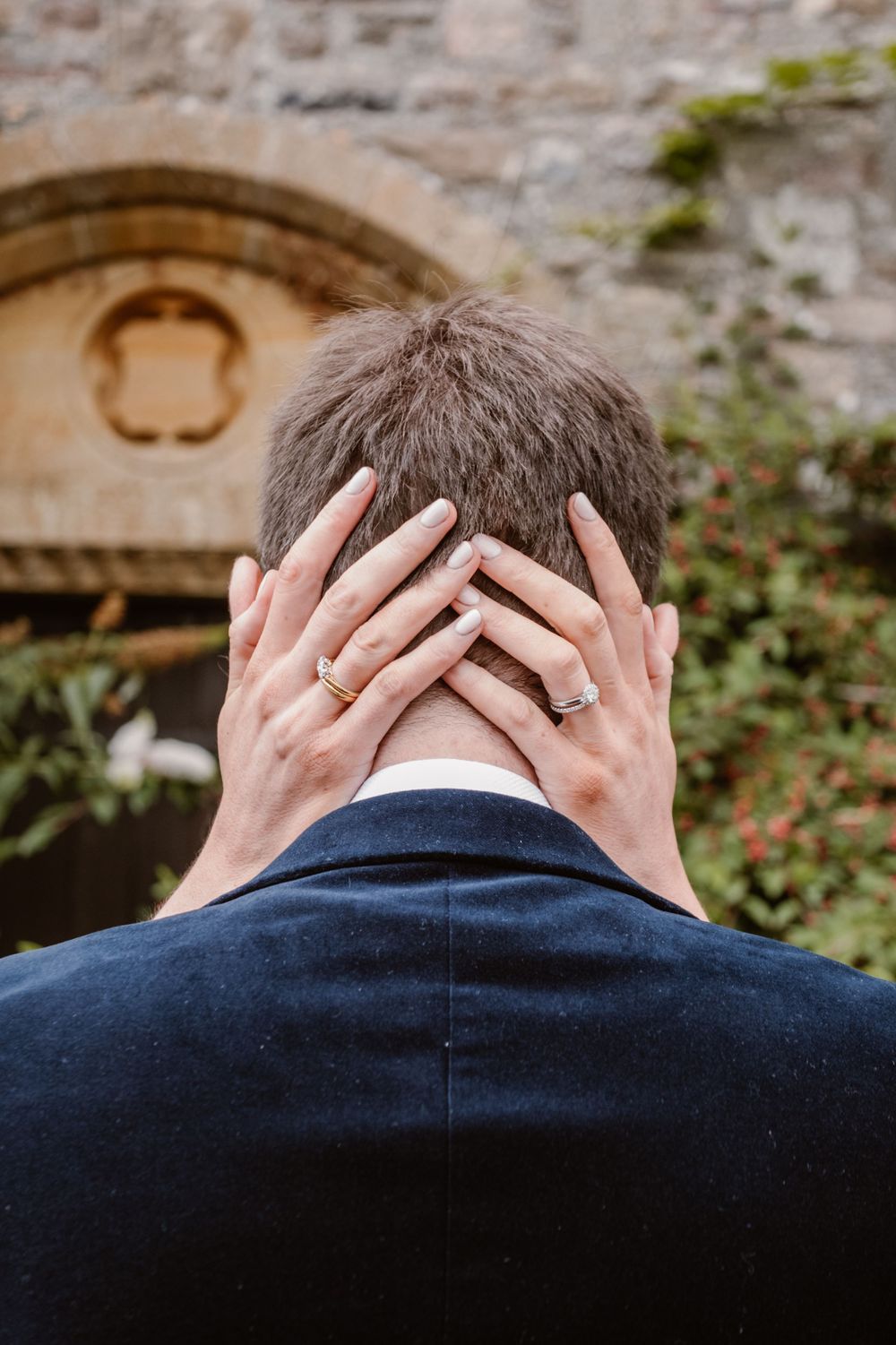 Newlywed couple embracing outdoors, showing wedding rings on hands placed on groom's head near stone building.