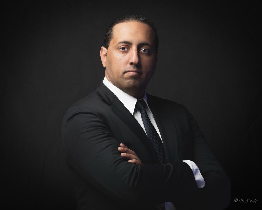 Professional man in black suit and tie with arms crossed posing against dark studio background.