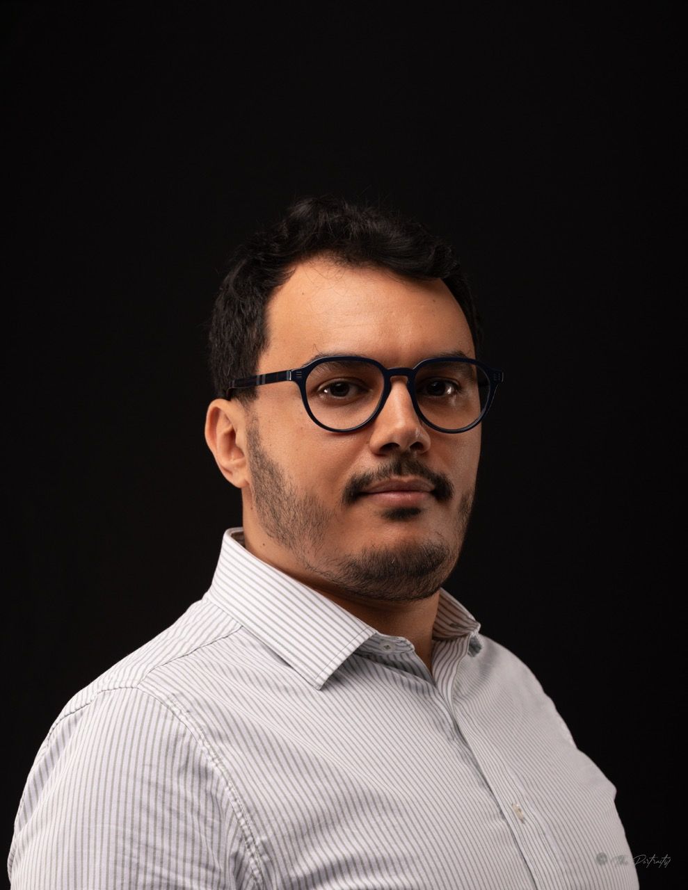 Young man with glasses and beard wearing striped shirt posing against black studio background.
