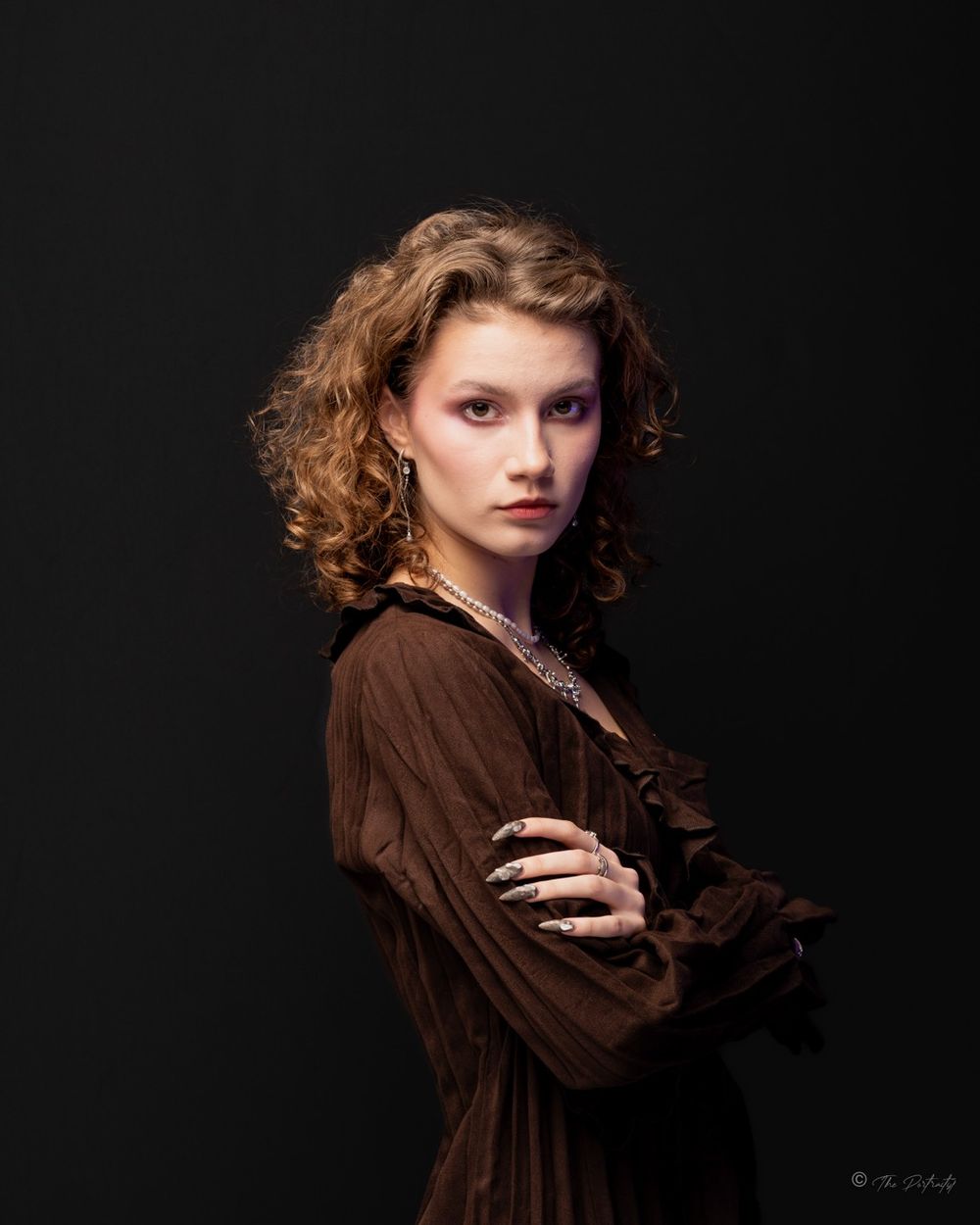 Young woman with curly hair in brown dress with arms crossed posing against dark background.