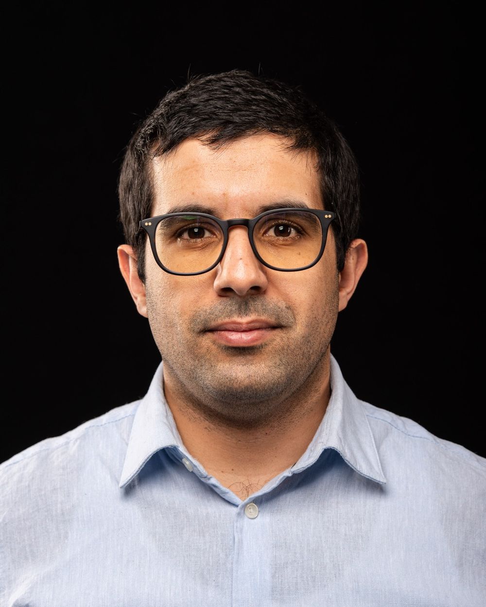 Man with glasses in light blue shirt against black background.