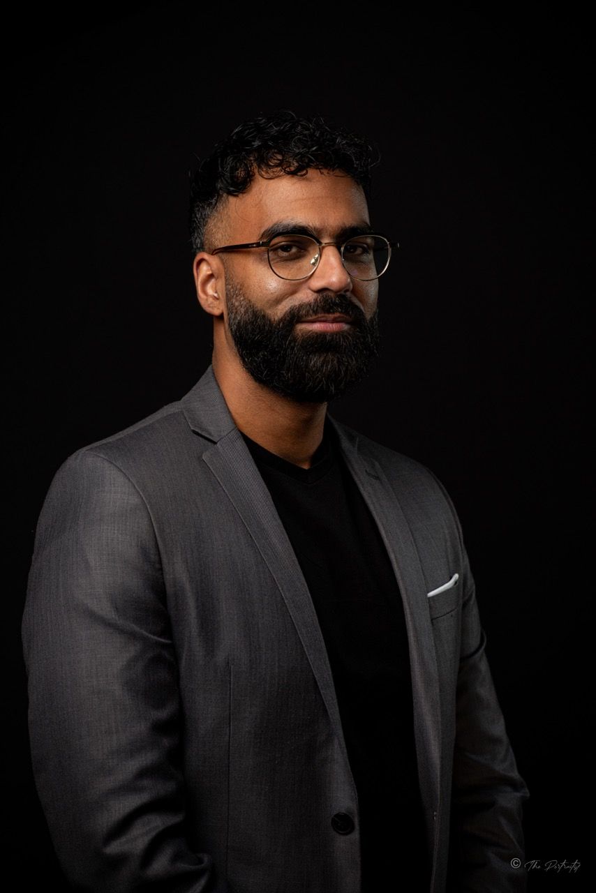 Bearded man with glasses in gray blazer against black background.
