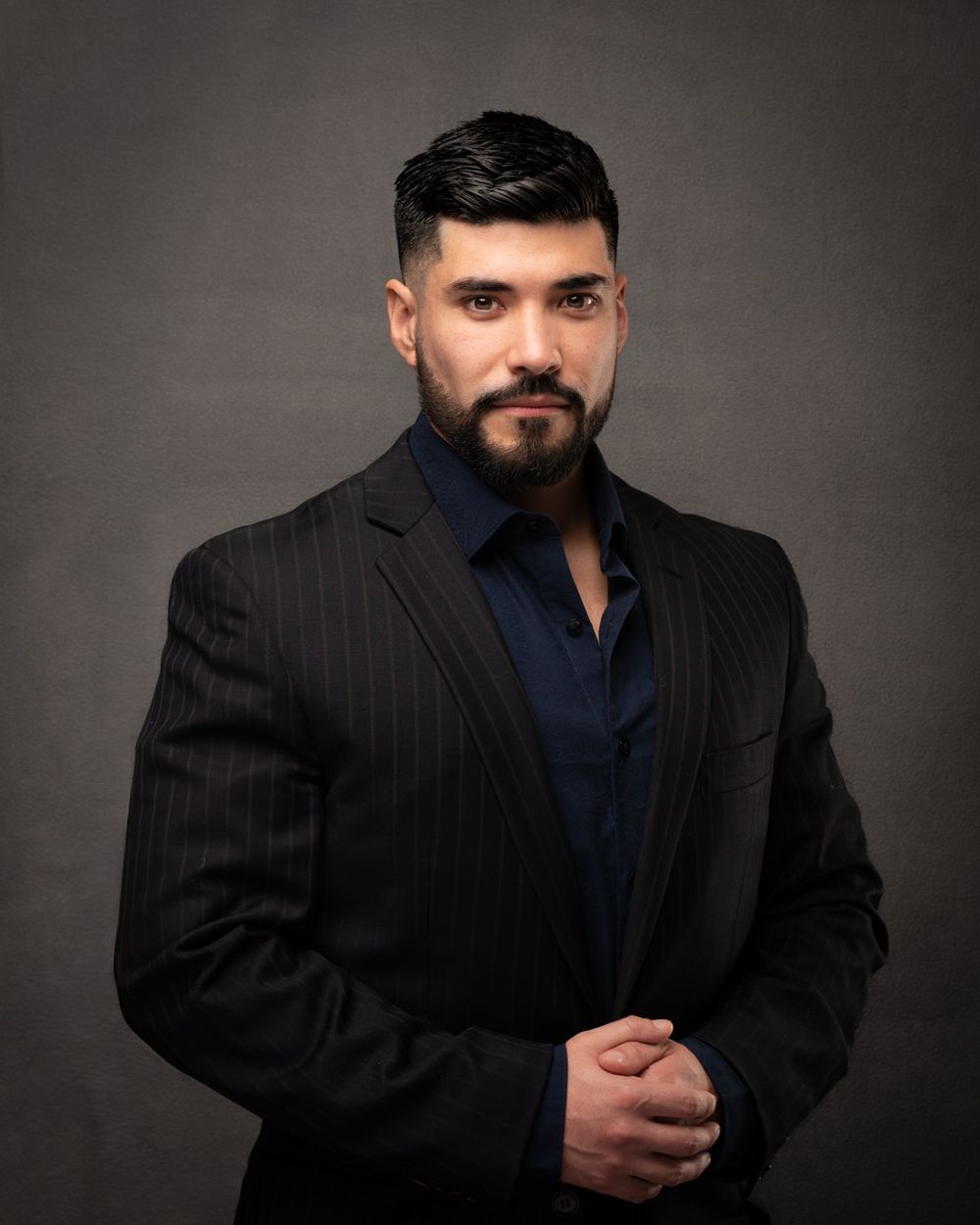 Man in black pinstripe suit with beard against gray background.