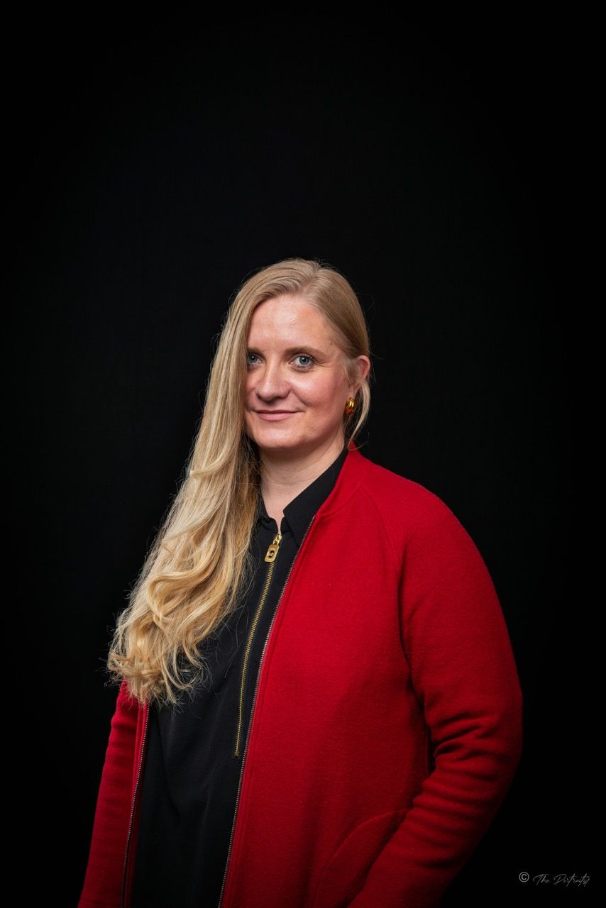 Woman in red jacket with blonde hair against black background.
