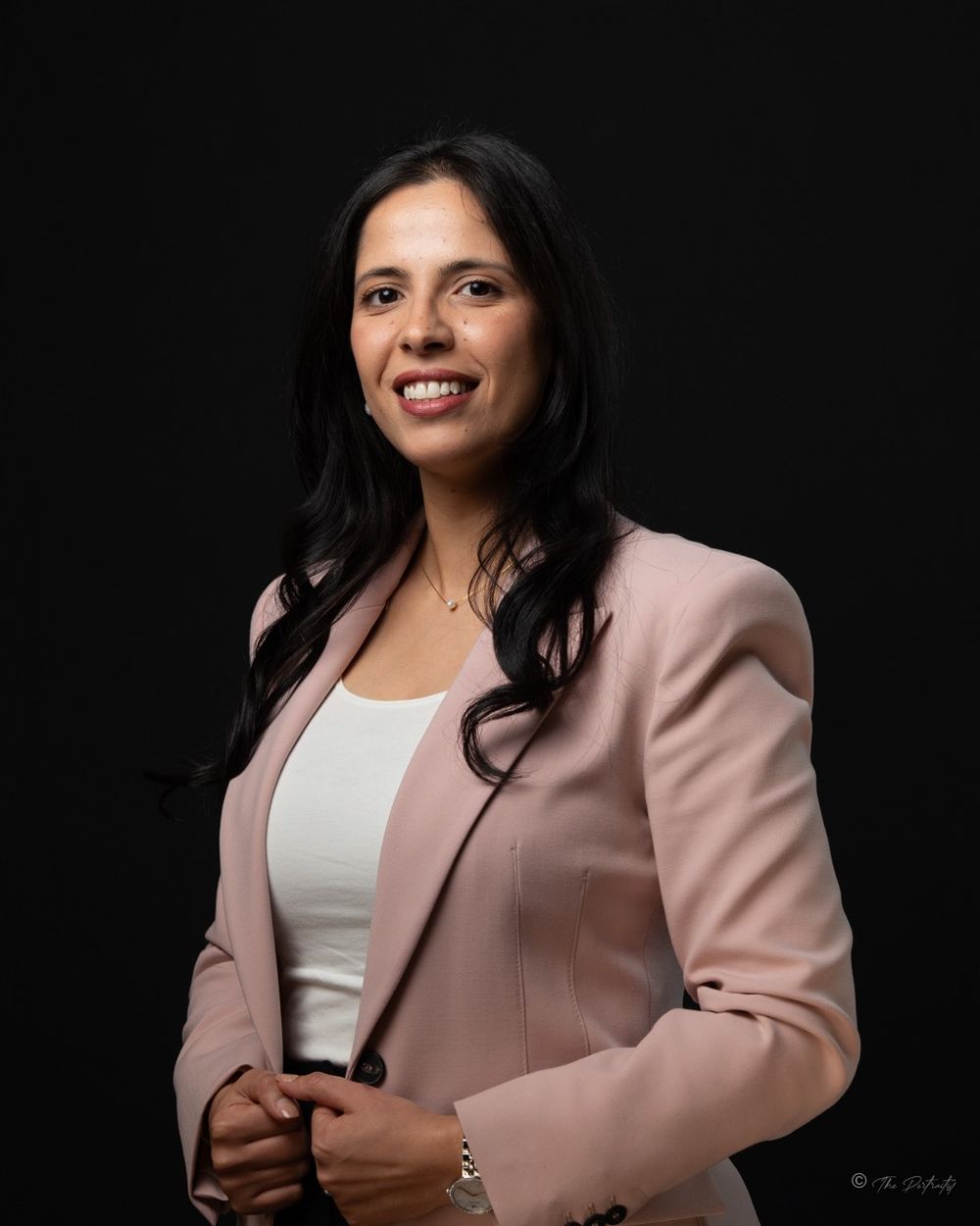 Professional woman in pink blazer smiles confidently against dark background in business portrait.