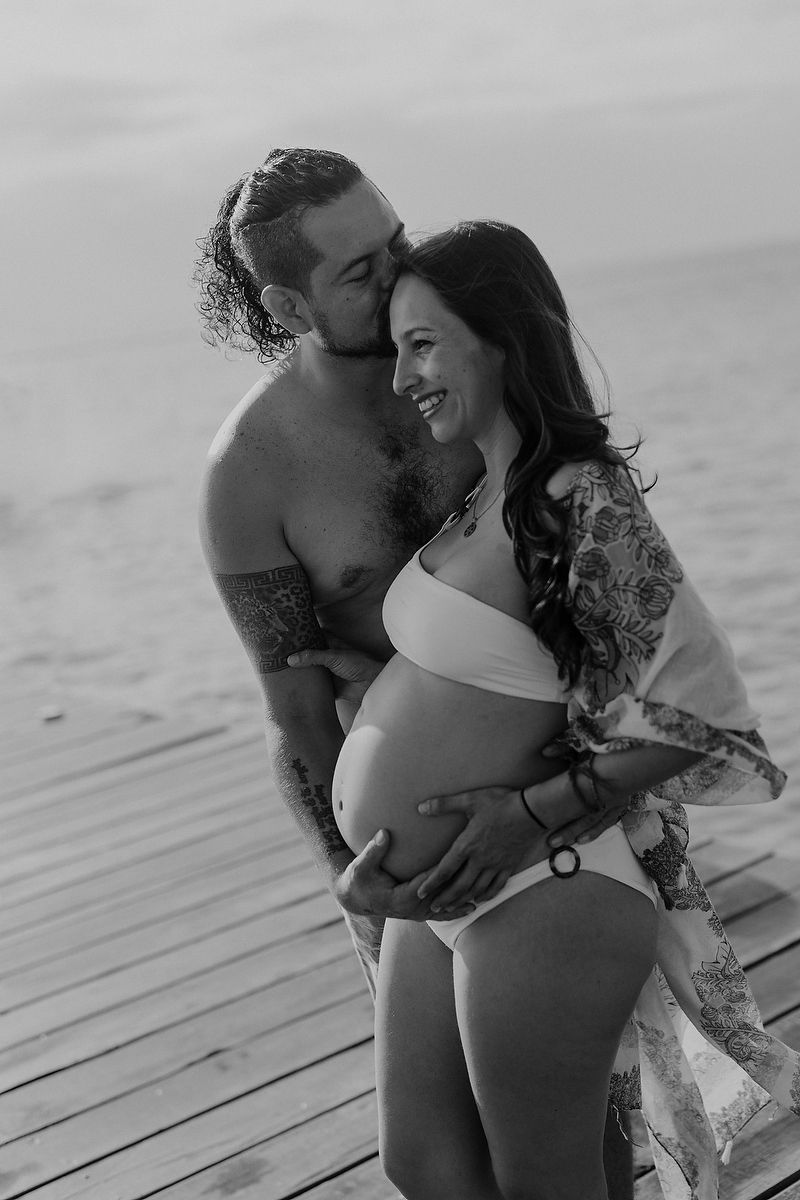 Black and white maternity photo of a couple on a wooden pier, man kissing pregnant womans cheek near the ocean.