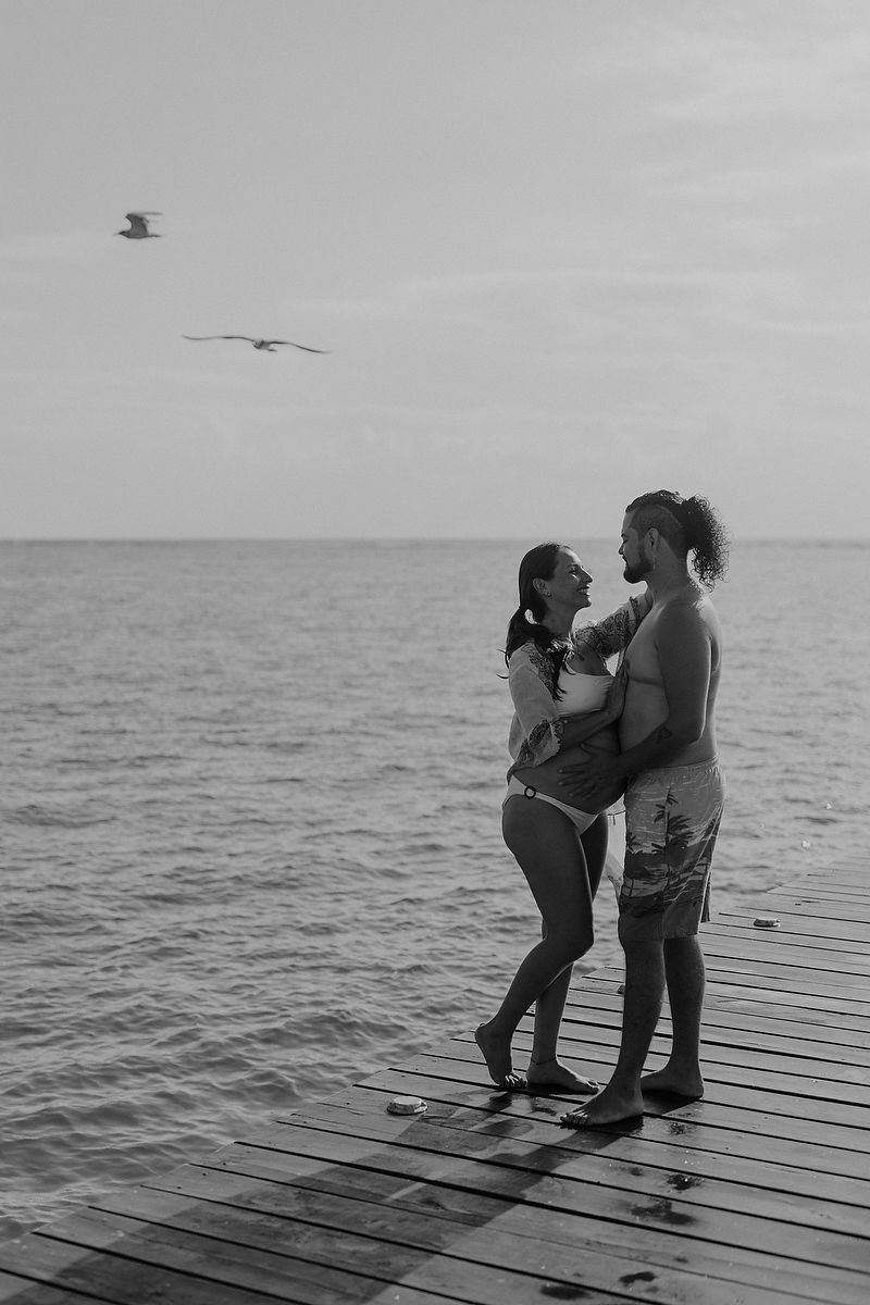 Black and white photo of a couple embracing on a wooden pier by the ocean with seagulls flying overhead.