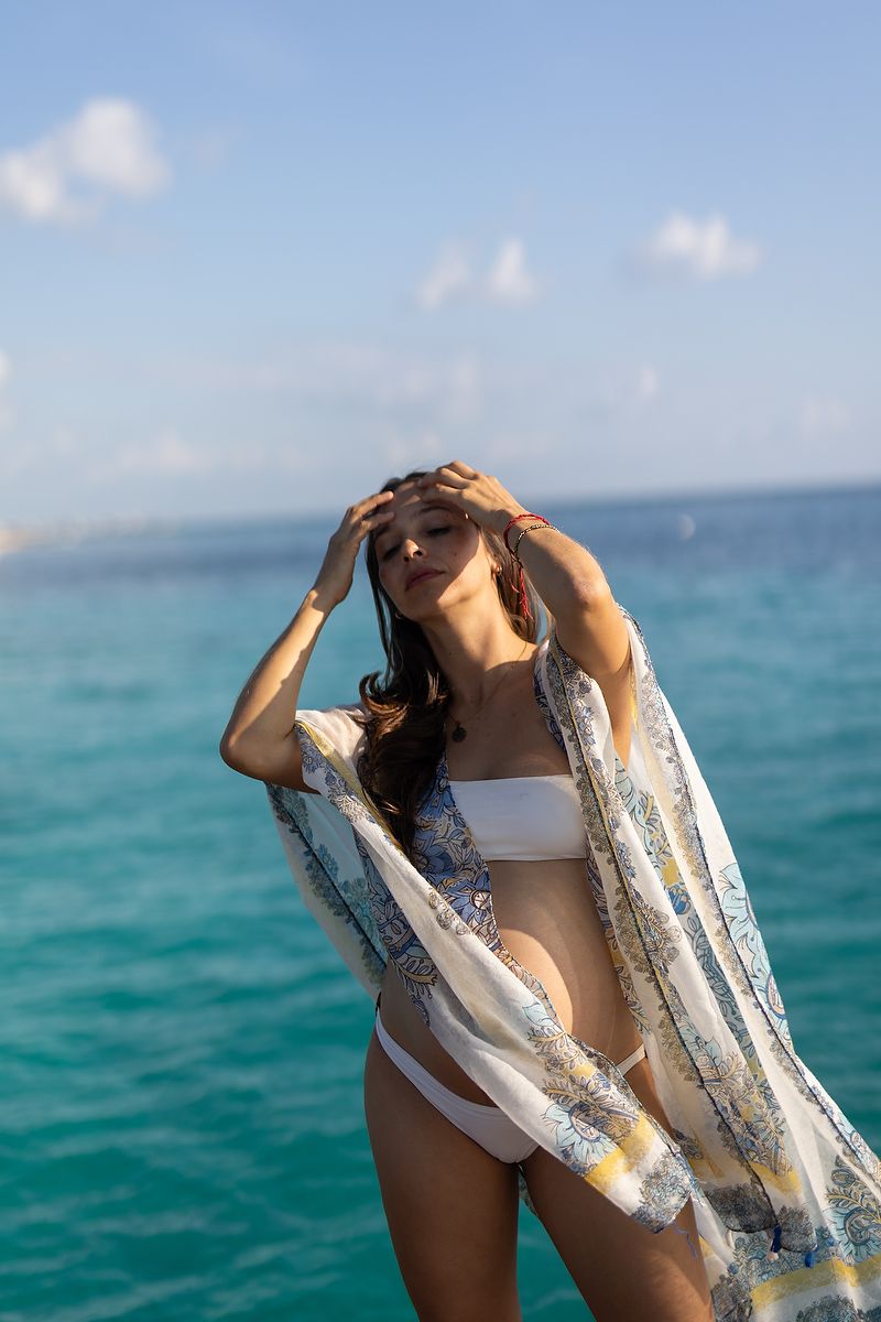 Woman in white bikini and floral kimono poses by turquoise ocean, hands raised to head on sunny day.