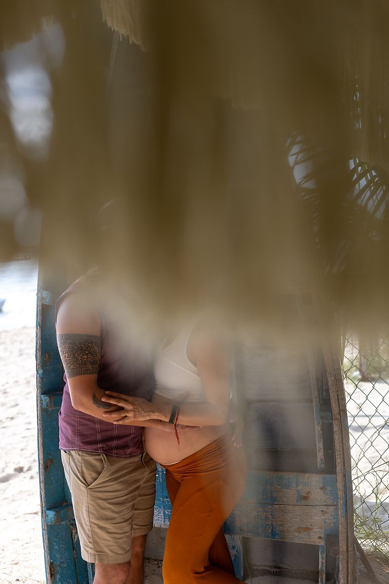 Couple embracing intimately outdoors, partially obscured by blurred foreground foliage near a weathered blue structure.
