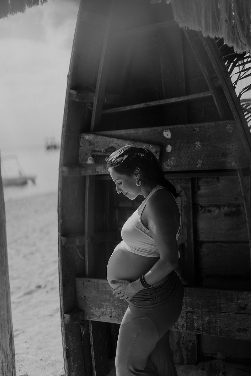 Black and white photo of a pregnant woman cradling her belly beside a wooden boat on a beach.