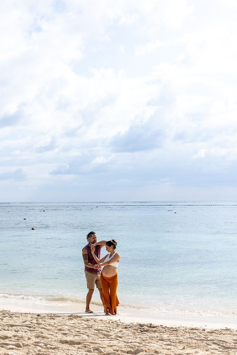 Happy expecting couple embracing on a white sand beach with calm turquoise ocean waters in the background.