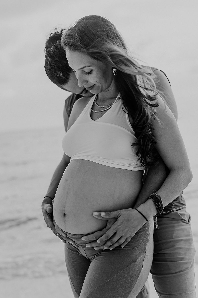 Black and white maternity photo of pregnant woman and partner embracing on beach, hands cradling baby bump.