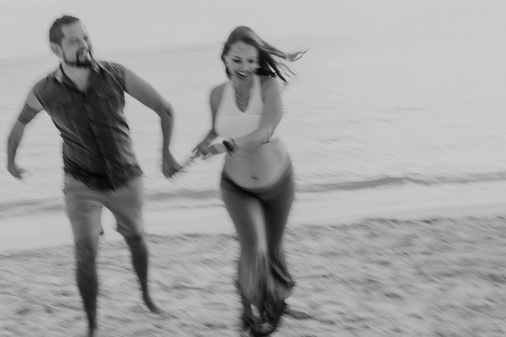 Black and white photo of a joyful couple running hand in hand along a beach shoreline.