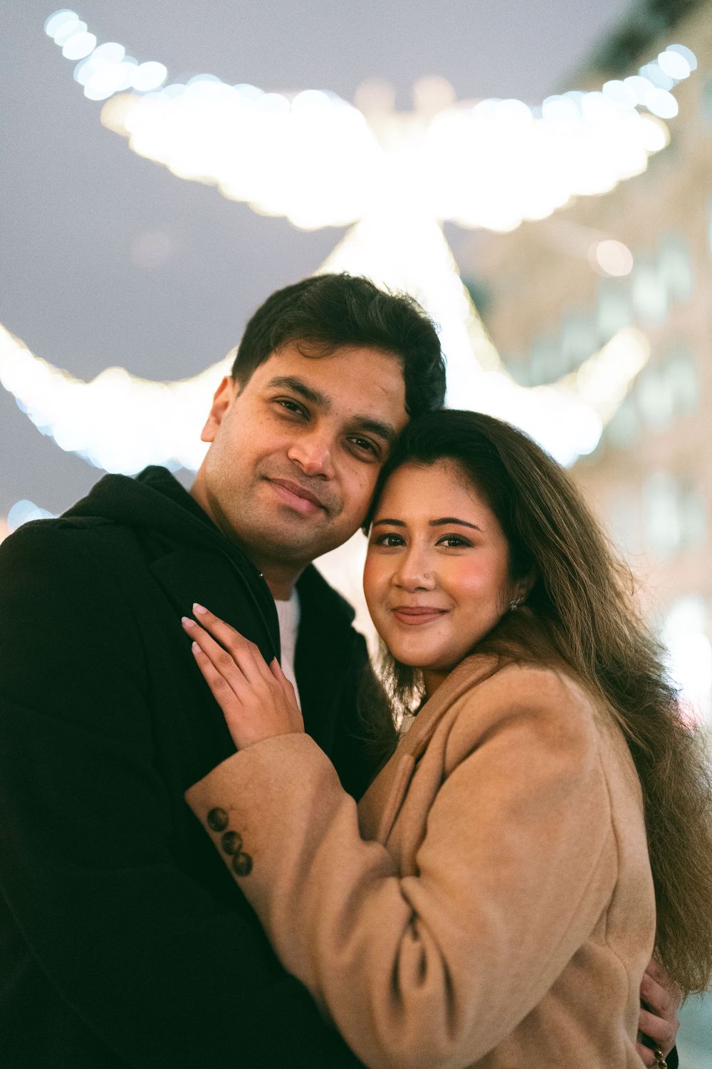 A couple embracing and smiling in front of bokeh holiday lights at night, woman in beige coat, man in black hoodie.