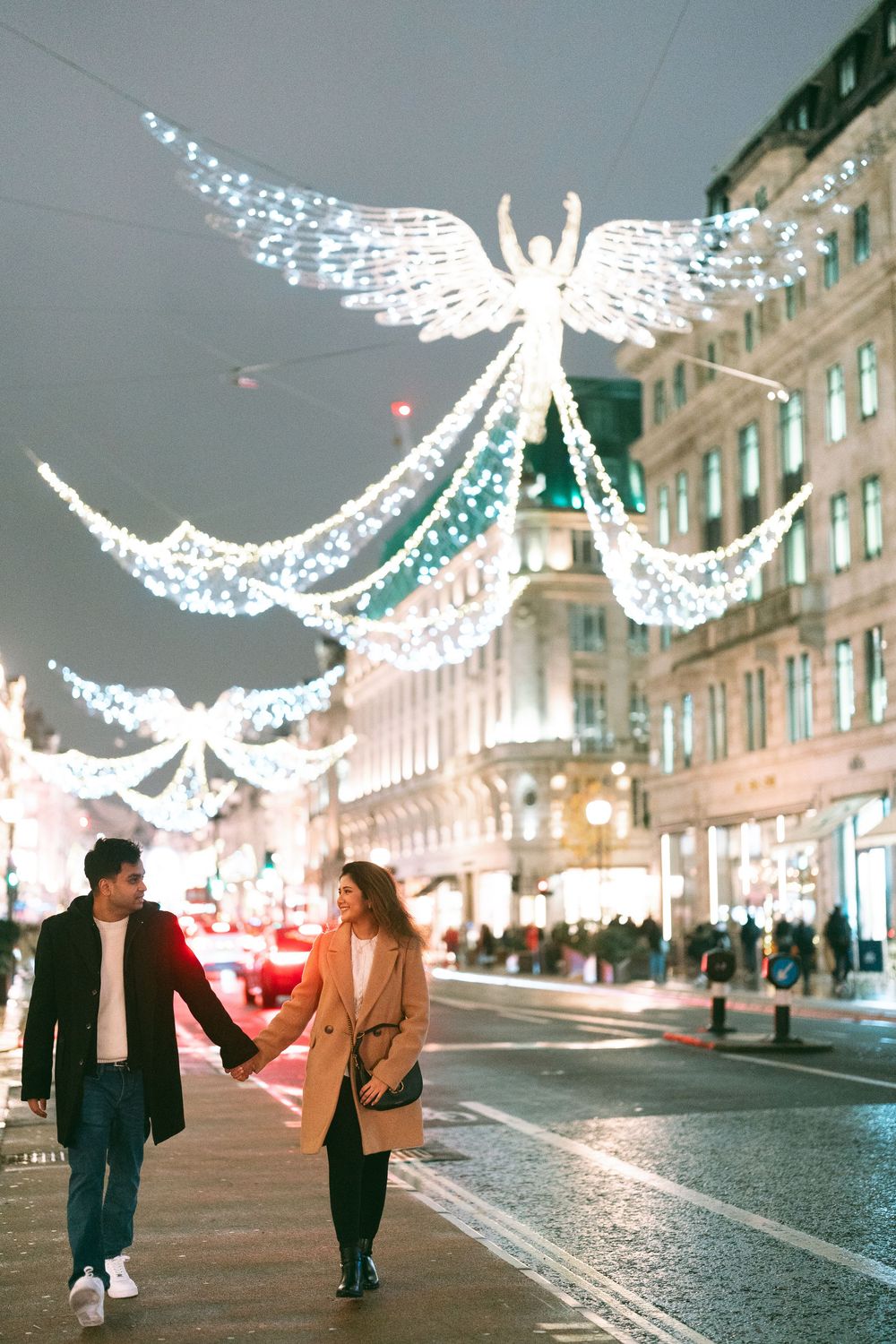 Couple holding hands walking on Regent Street London at night with festive angel Christmas lights illuminating the street.