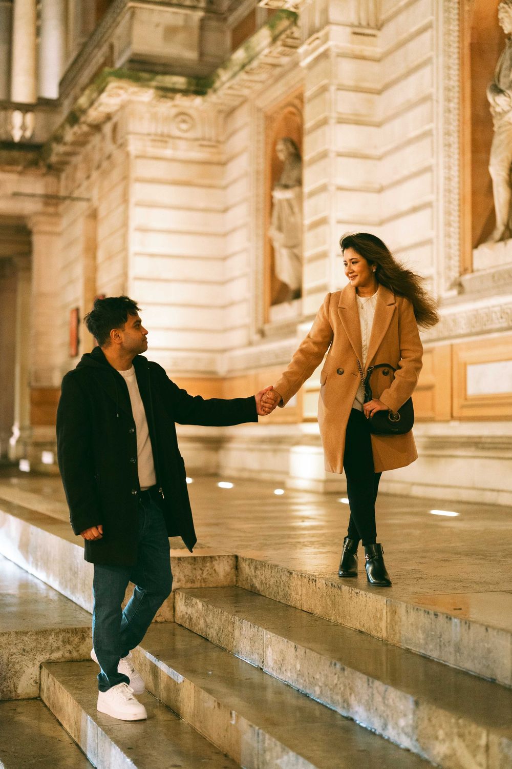 Couple holding hands on illuminated stone steps outside a classical building at night, woman in camel coat smiling.