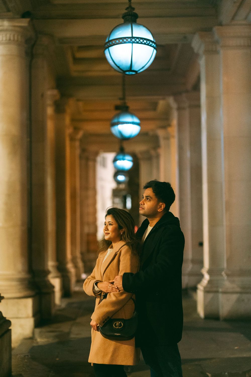 Couple standing under glowing blue lanterns in an elegant columned arcade at night, woman in beige coat, man in black.