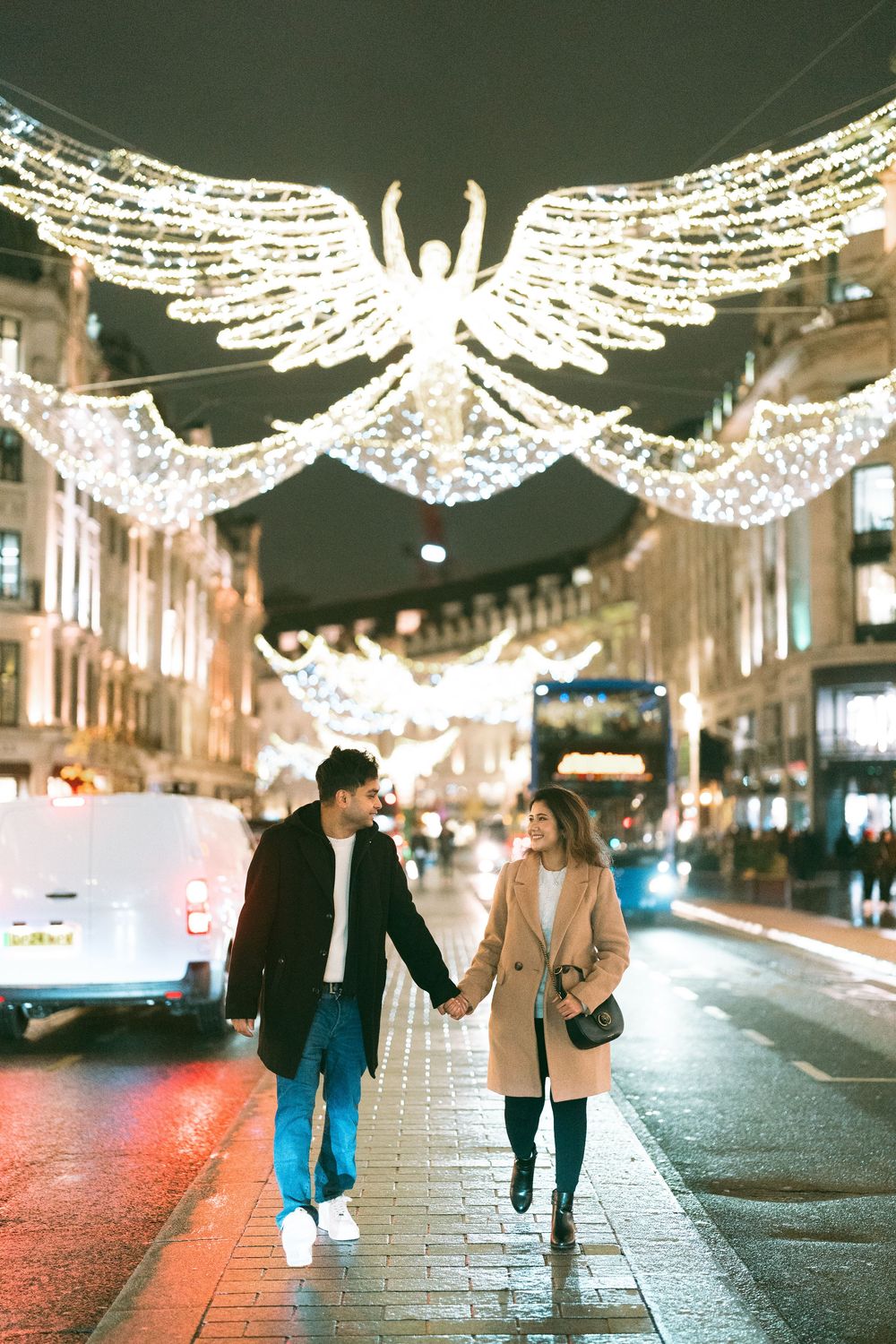 Couple holding hands on Regent Street London at night with glowing angel Christmas lights overhead.