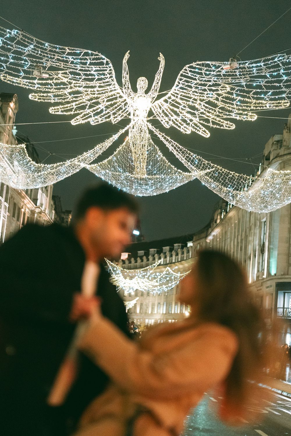 Couple sharing a romantic moment under glowing angel Christmas lights on a festive city street at night.