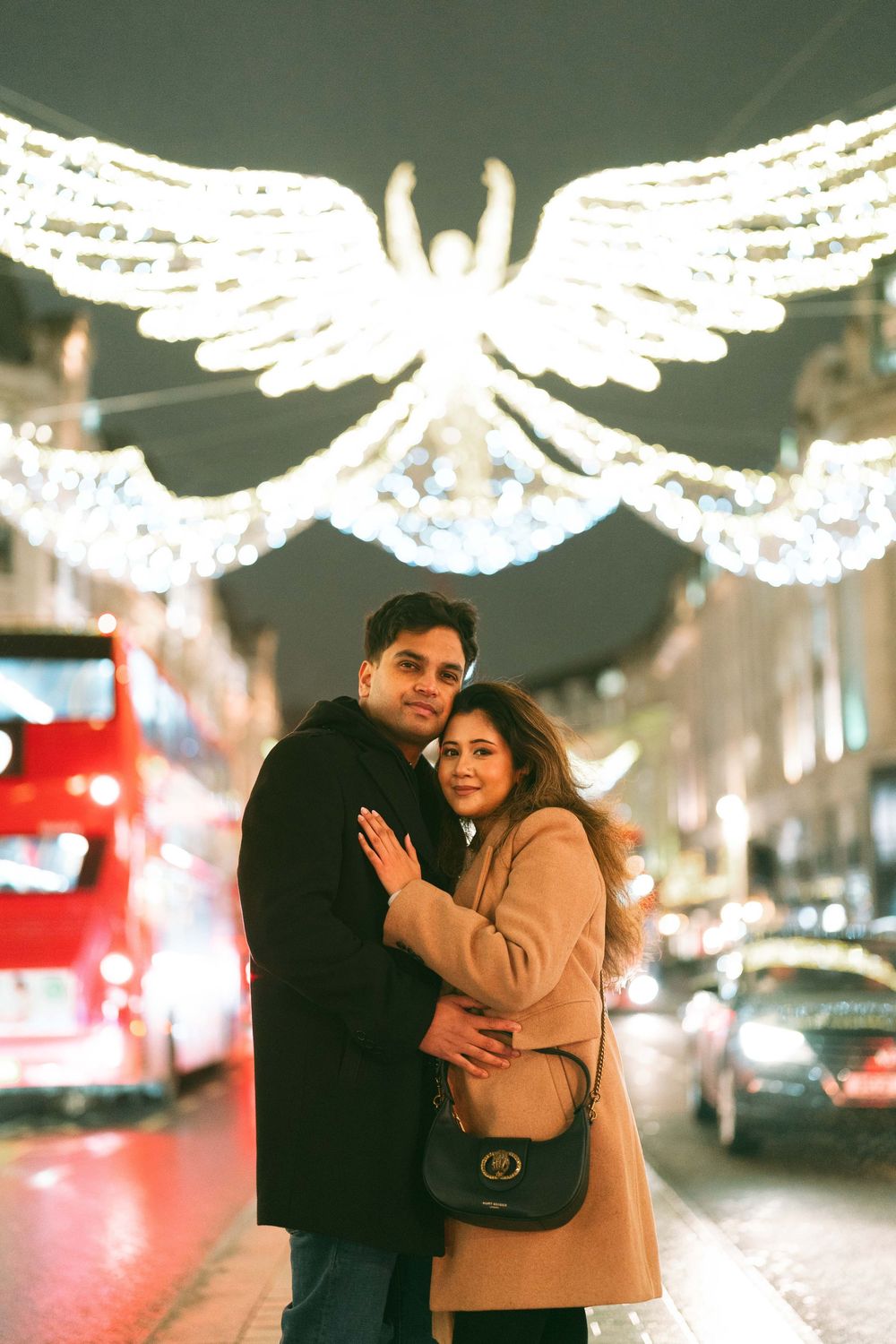 Couple embracing on a city street at night with glowing angel wing Christmas lights overhead.