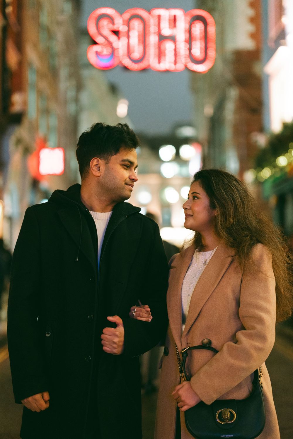 Couple standing together at night in Soho, London, with neon signs and city lights in the background.