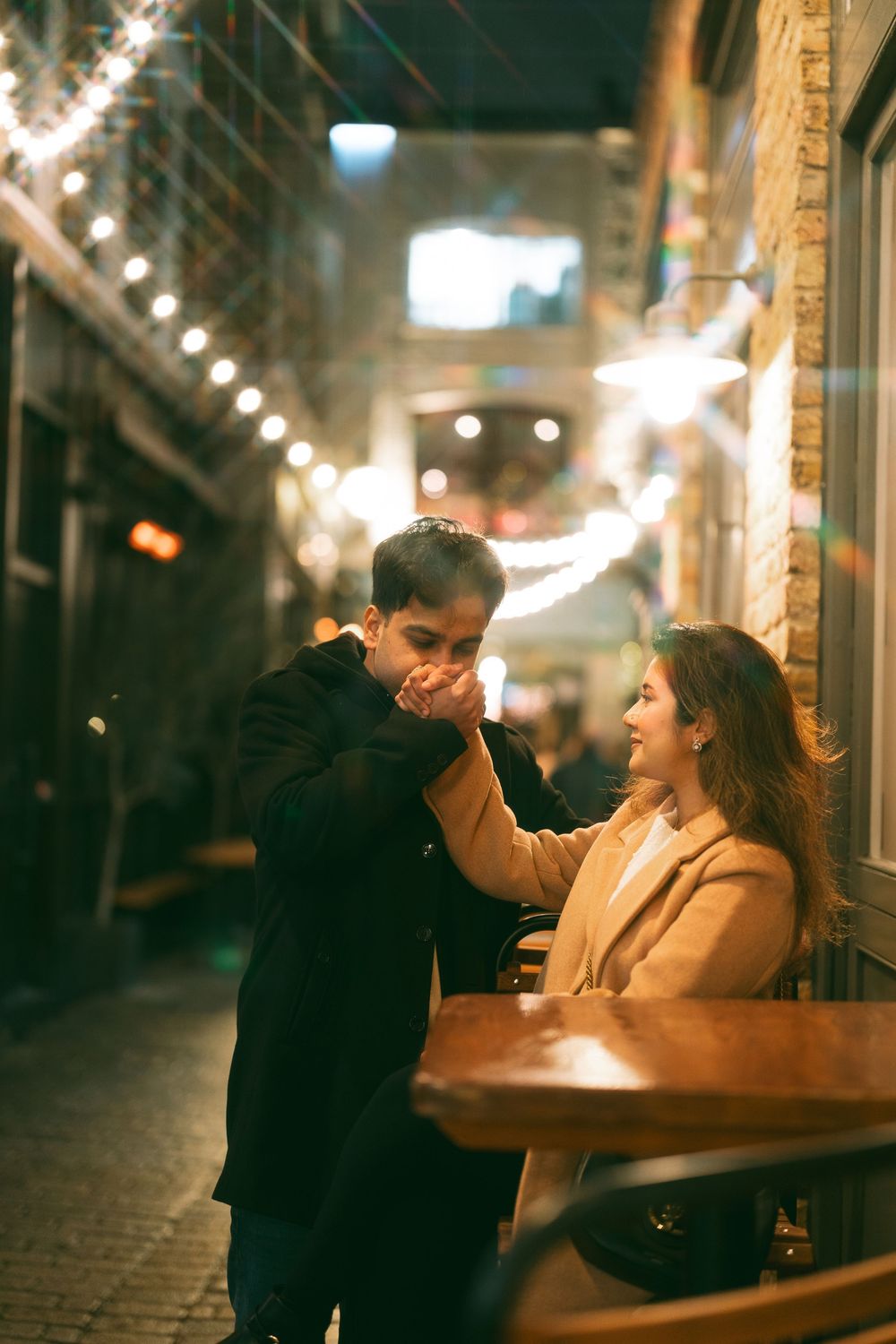 Romantic couple on a date at night, man kissing woman's hand at outdoor cafe with warm string lights.