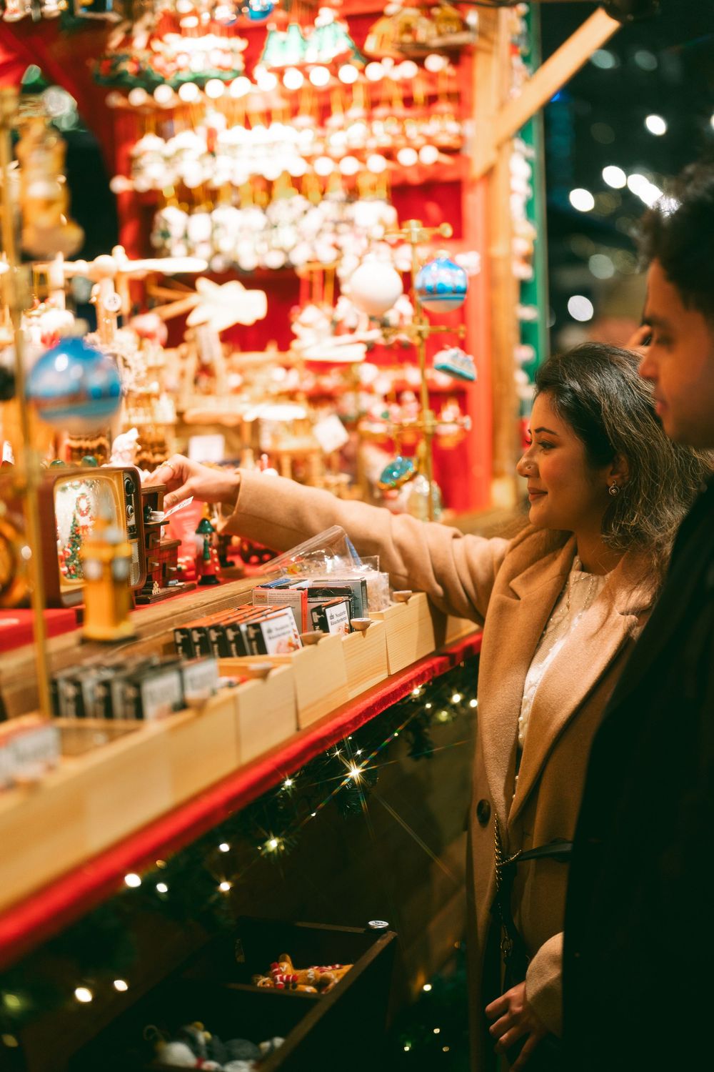 Couple browsing colorful Christmas market stall with ornaments and festive decorations at night.