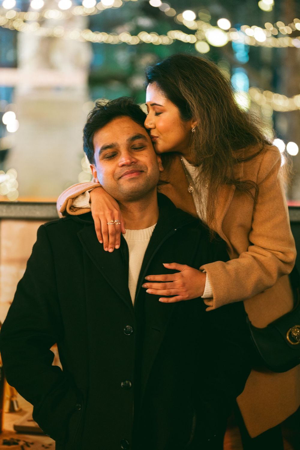 Romantic couple embracing outdoors at night, woman kissing man on cheek with warm bokeh lights in background.