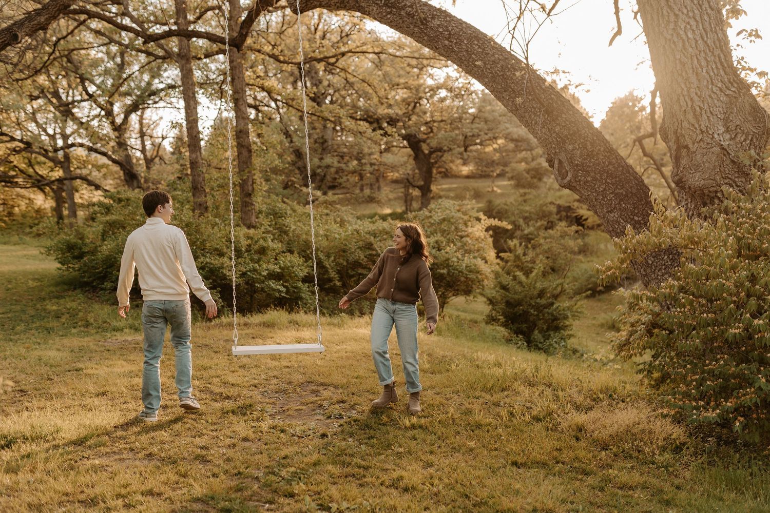Couple walking toward a wooden tree swing in a sunlit green meadow beneath a large oak tree.