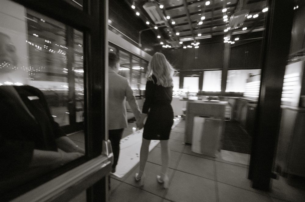 Black and white photo of a couple walking hand in hand through a modern building entrance at night.