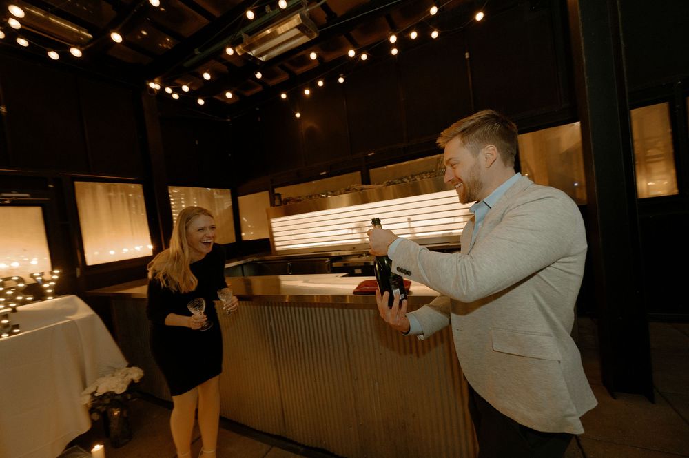 Man in blazer opening wine bottle for woman in black dress at dimly lit restaurant with string lights.