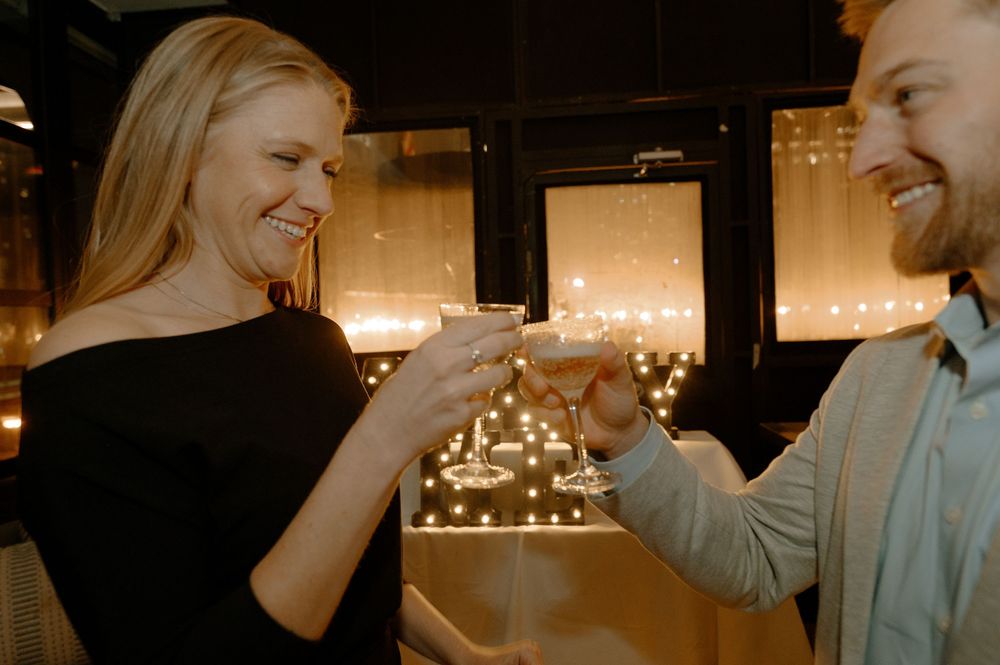 A smiling couple toasting with cocktails by candlelight in a romantic restaurant setting.