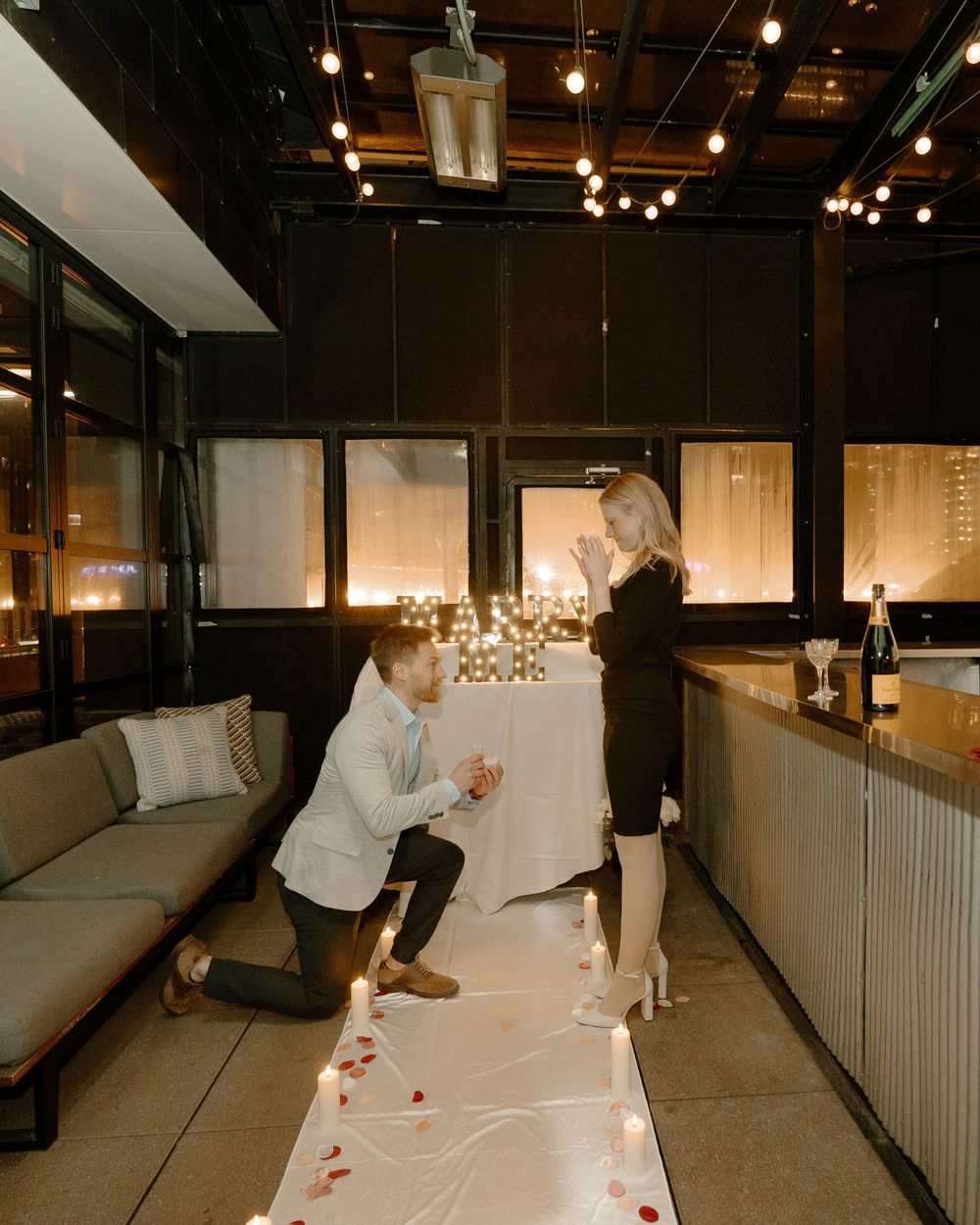 Man proposes on one knee to woman in romantic candlelit venue with rose petals and string lights.