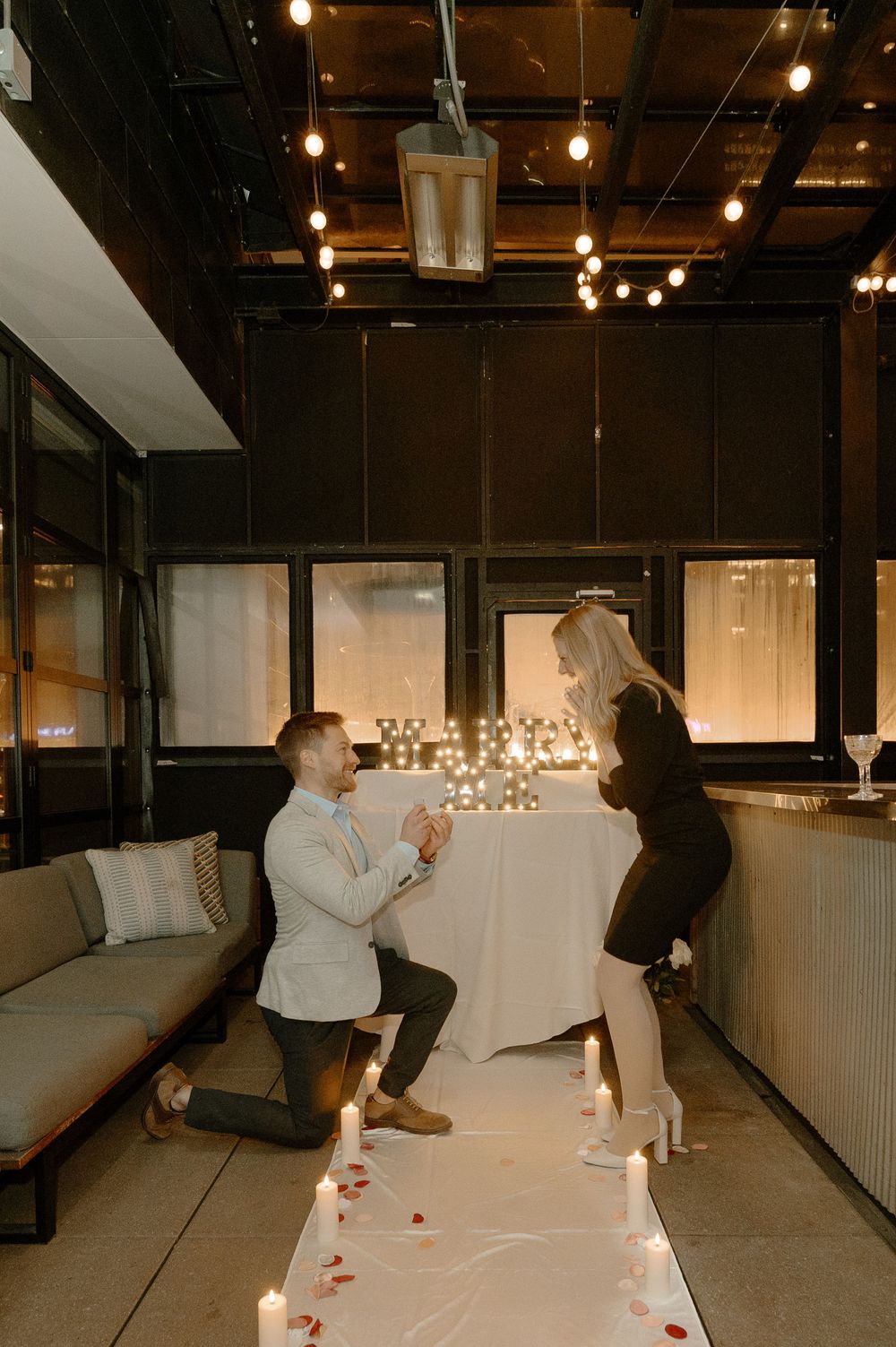 Man proposes on one knee to woman in romantic candlelit venue with string lights and marquee letters.
