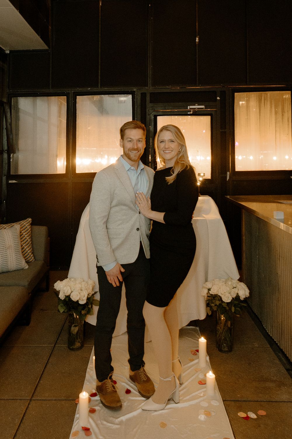 Couple smiling together surrounded by candles and white roses during a romantic proposal setup indoors.