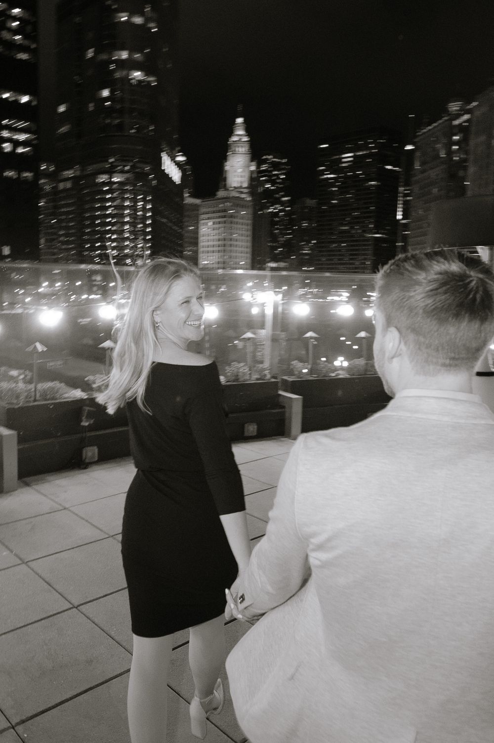 Black and white photo of a couple on a rooftop terrace at night with illuminated city skyline in background.