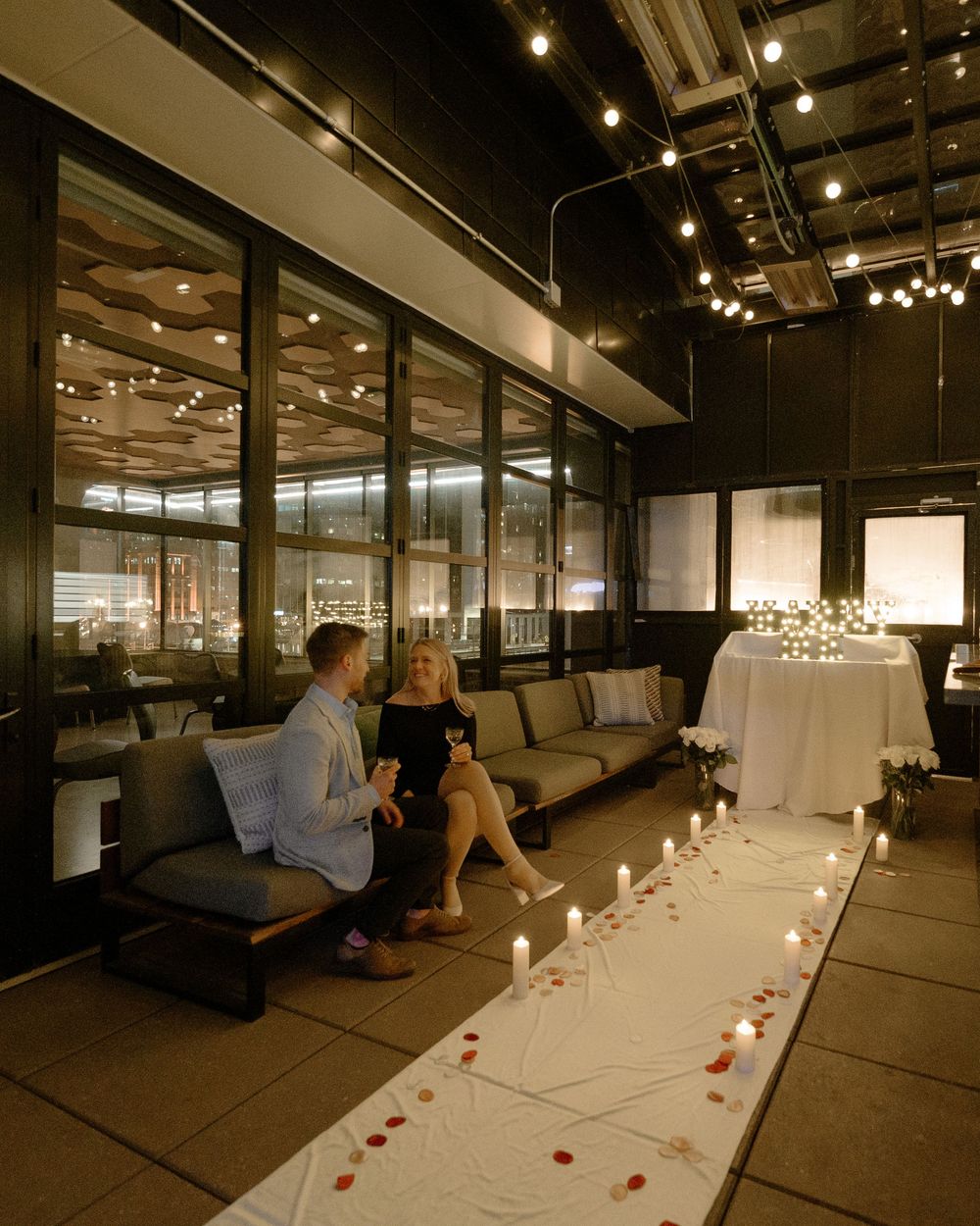 Romantic candlelit proposal setup with rose petals leading to a decorated table while couple sits together.