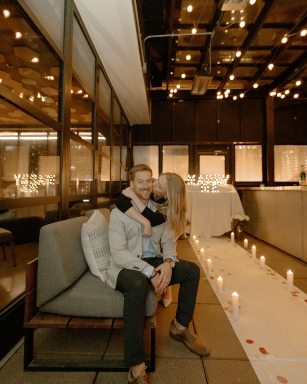 Couple sitting on a sofa surrounded by candles and string lights in a romantic indoor venue.
