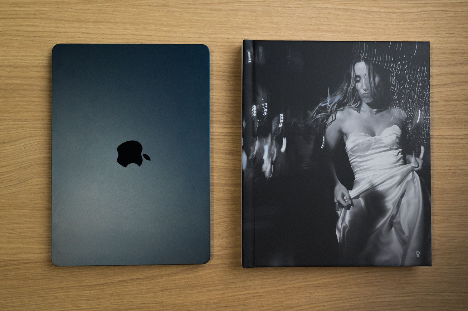 Black hardcover photography book beside an Apple MacBook on a wooden surface.