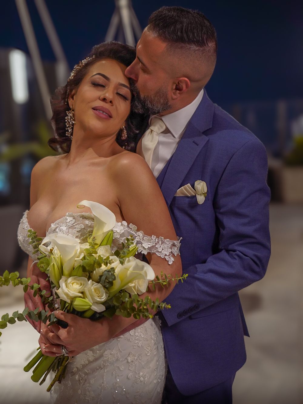 Bride and groom share romantic wedding moment, she holds white floral bouquet while he kisses her cheek at night.