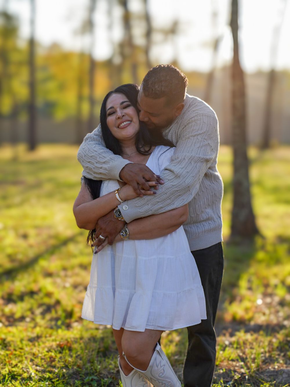 Happy couple hugging and laughing outdoors in golden sunlight, woman in white dress and cowboy boots.