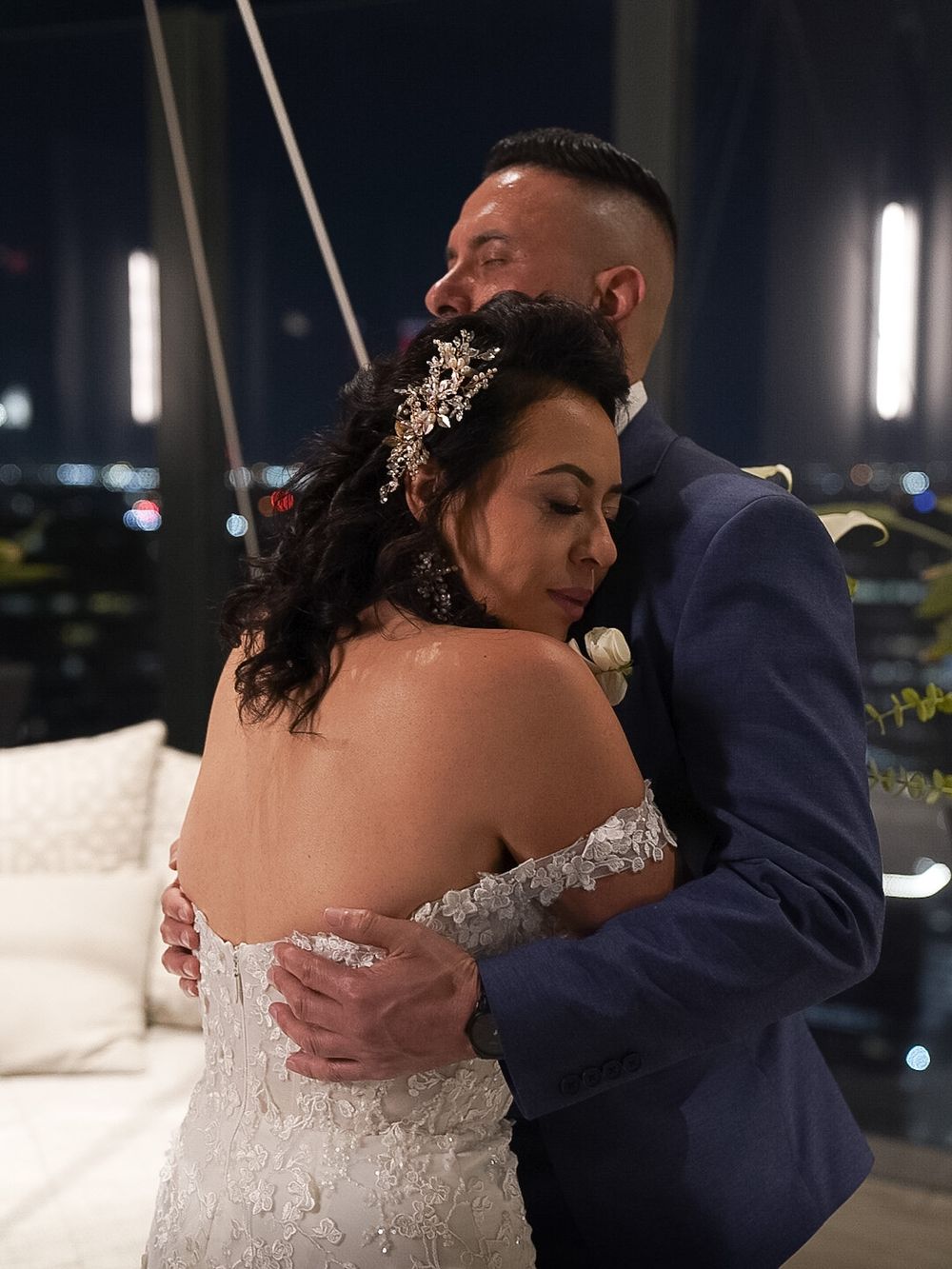 A bride and groom share a romantic embrace on a rooftop at night, with city lights glowing in the background.
