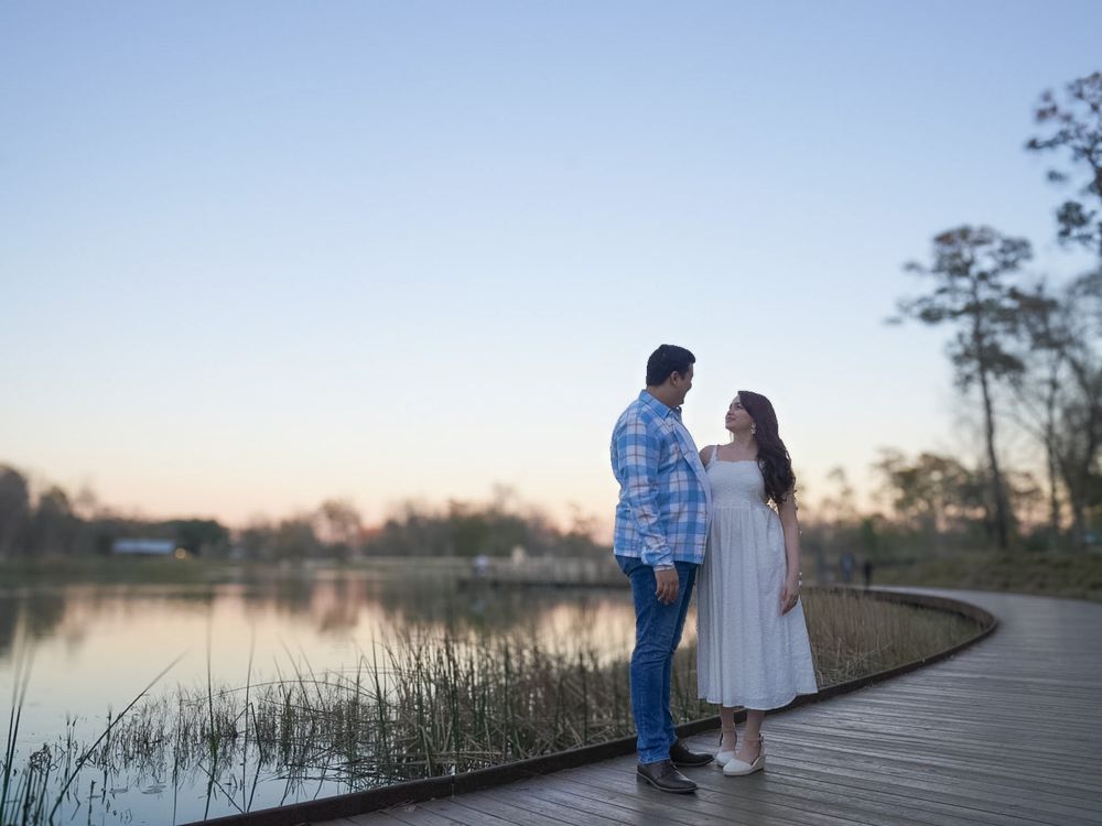 Couple standing on a wooden dock by a serene lake at sunset, surrounded by tall grasses and trees.