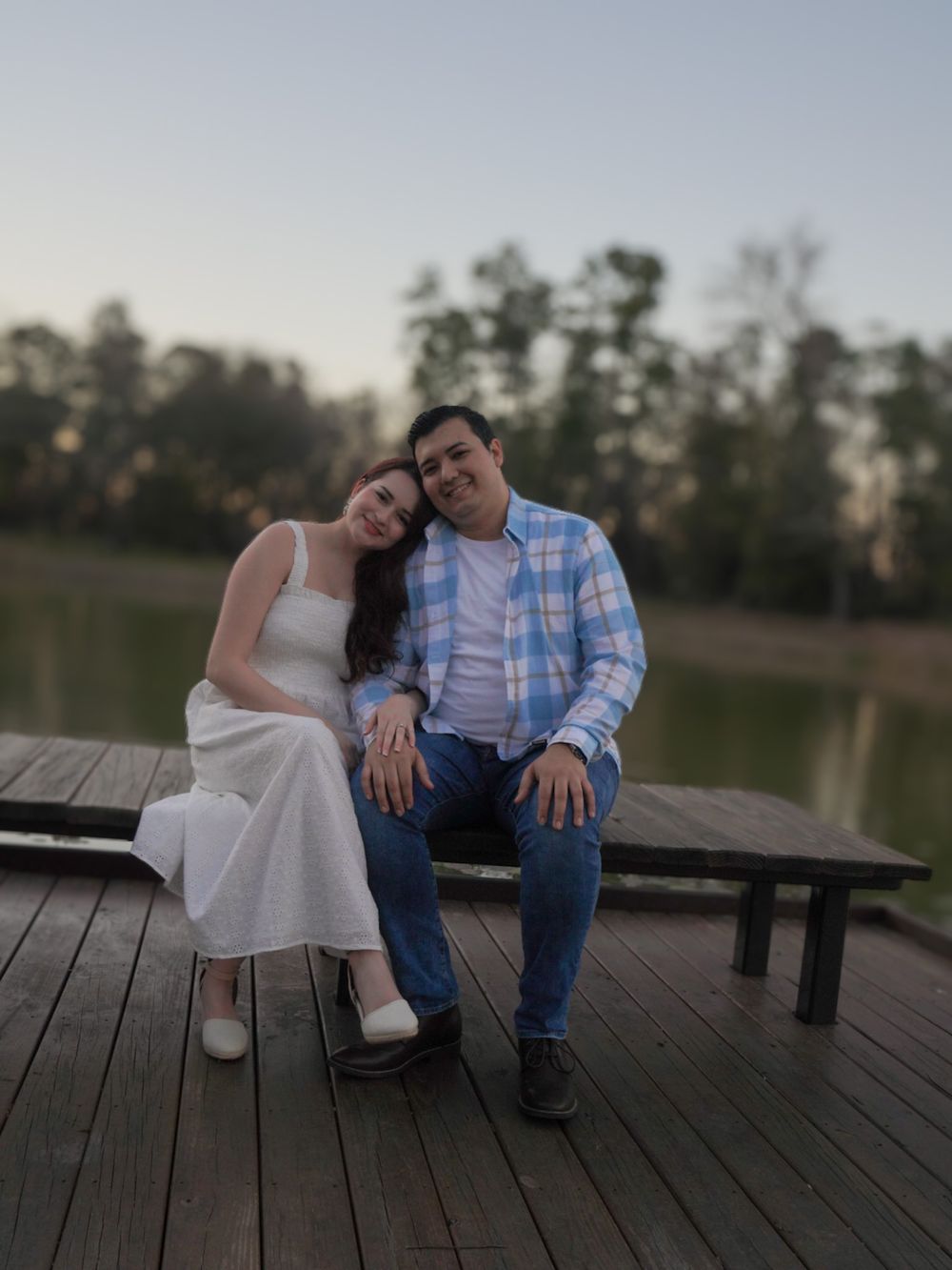 Couple sitting together on a wooden dock by a lake at dusk, woman in white dress, man in plaid shirt and jeans.