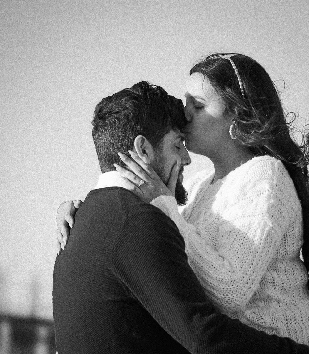 Black and white photo of a couple sharing a romantic kiss outdoors, woman in white sweater embracing man in dark jacket.